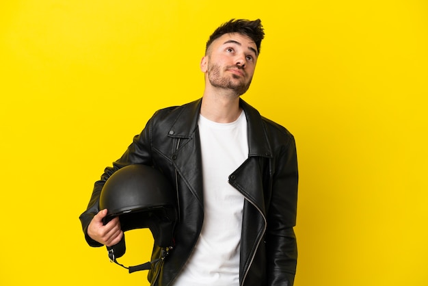 Young caucasian man with a motorcycle helmet isolated on yellow background and looking up