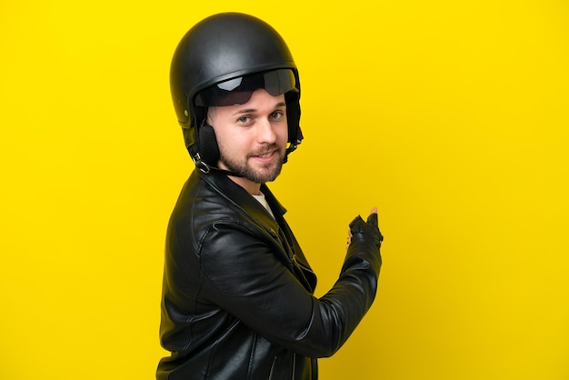 Young caucasian man with a motorcycle helmet isolated on yellow background pointing back