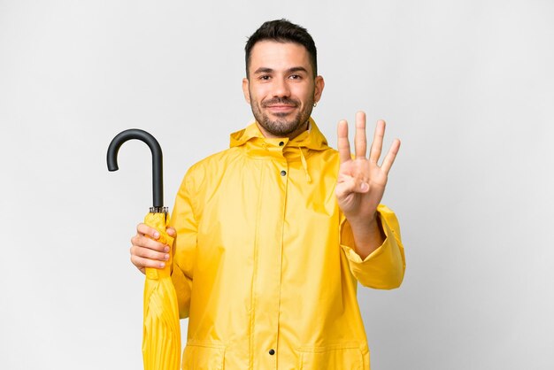 Young caucasian man with rainproof coat and umbrella over isolated white background happy and counting four with fingers