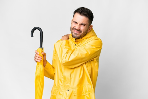 努力をしたために肩の痛みに苦しんでいる孤立した白い背景の上に防雨コートと傘を持つ若い白人男性