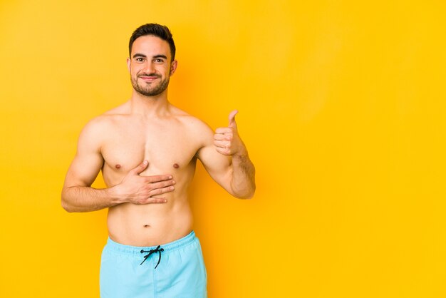 Young caucasian man with swimsuit isolated on yellow wall Young caucasian man with trtouches tummy, smiles gently, eating and satisfaction concept.