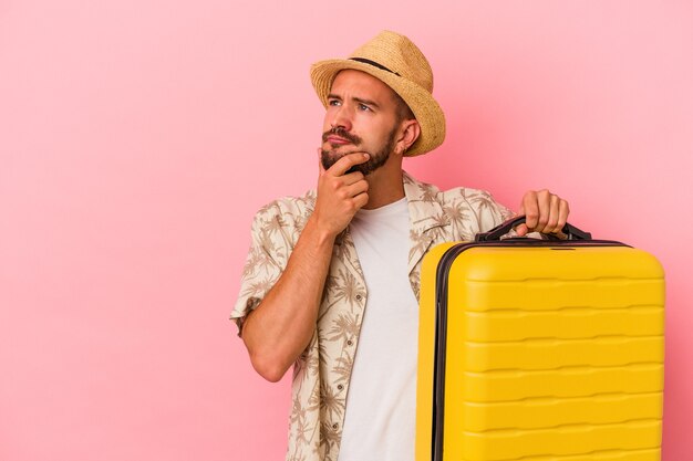 Young caucasian man with tattoos going to travel isolated on pink background  looking sideways with doubtful and skeptical expression.