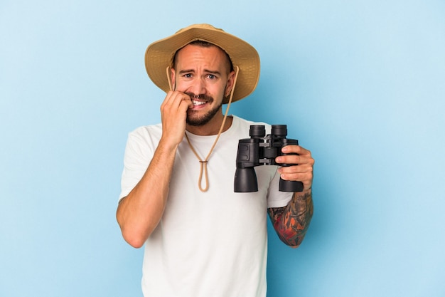 Young caucasian man with tattoos holding binoculars isolated on blue background  biting fingernails, nervous and very anxious.