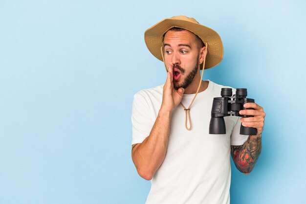 Young caucasian man with tattoos holding binoculars isolated on blue background  is saying a secret hot braking news and looking aside