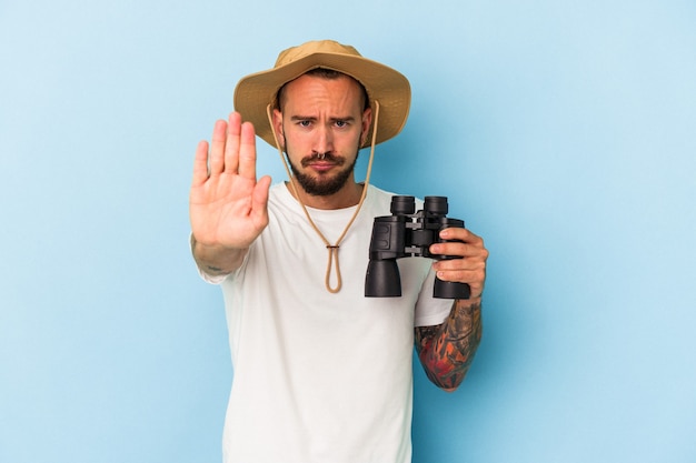 Young caucasian man with tattoos holding binoculars isolated on blue background  standing with outstretched hand showing stop sign, preventing you.