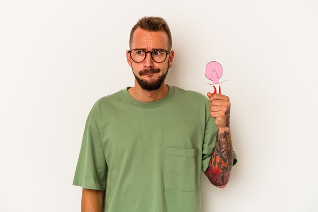 Young caucasian man with tattoos holding lollipop isolated on white background  confused, feels doubtful and unsure.
