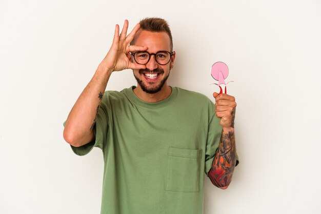 Young caucasian man with tattoos holding lollipop isolated on white background  excited keeping ok gesture on eye.