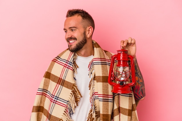 Young caucasian man with tattoos holding vintage lantern isolated on pink background  looks aside smiling, cheerful and pleasant.