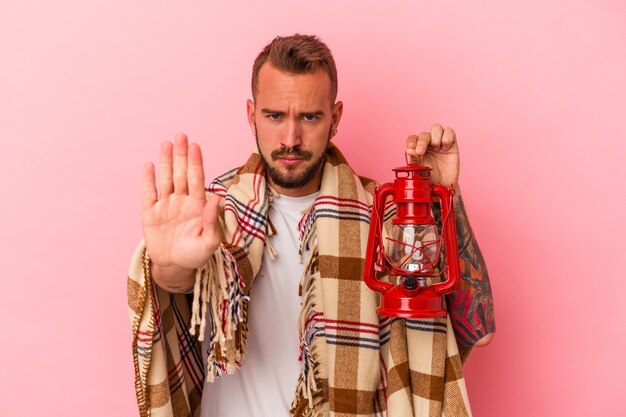 Young caucasian man with tattoos holding vintage lantern isolated on pink background  standing with outstretched hand showing stop sign, preventing you.