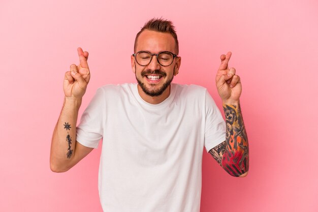 Young caucasian man with tattoos isolated on pink background  crossing fingers for having luck