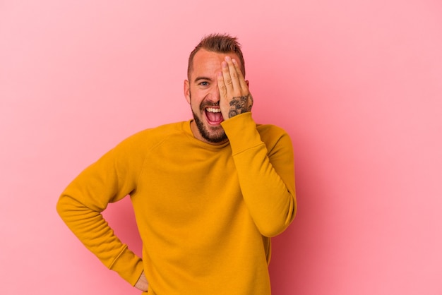 Young caucasian man with tattoos isolated on pink background  having fun covering half of face with palm.