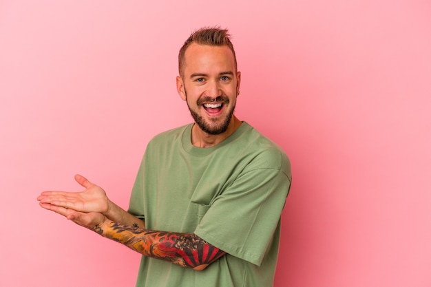 Young caucasian man with tattoos isolated on pink background  holding a copy space on a palm.