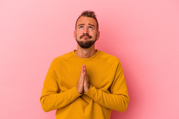 Young caucasian man with tattoos isolated on pink background  holding hands in pray near mouth, feels confident.