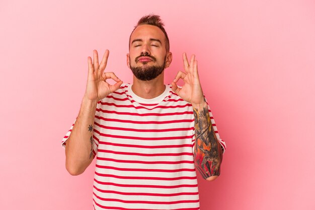 Young caucasian man with tattoos isolated on pink background  relaxes after hard working day, she is performing yoga.