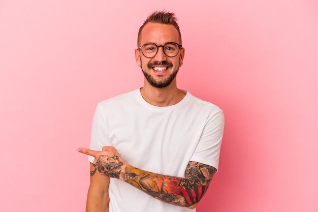 Young caucasian man with tattoos isolated on pink background  smiling and pointing aside, showing something at blank space.