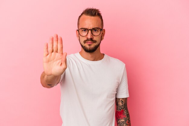 Young caucasian man with tattoos isolated on pink background  standing with outstretched hand showing stop sign, preventing you.