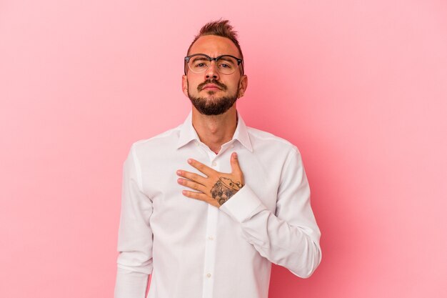 Young caucasian man with tattoos isolated on pink background taking an oath, putting hand on chest