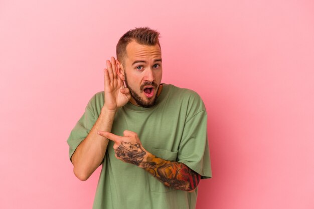 Young caucasian man with tattoos isolated on pink background  trying to listening a gossip.