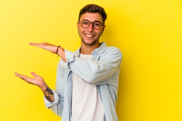 Young caucasian man with tattoos isolated on yellow background  holding something little with forefingers, smiling and confident.