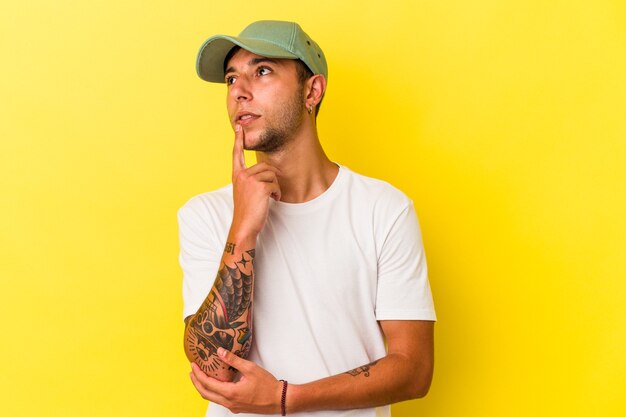 Young caucasian man with tattoos isolated on yellow background  looking sideways with doubtful and skeptical expression.