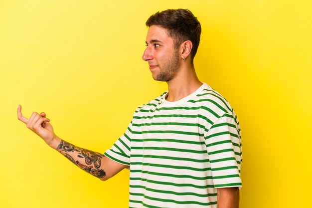 Молодой кавказский мужчина с татуировками, изолированными на желтом фоне, указывая пальцем на вас, как будто приглашая подойти ближе.