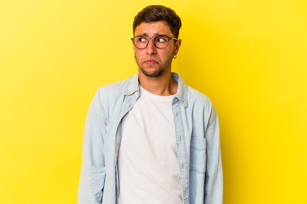 Young caucasian man with tattoos isolated on yellow background  shrugs shoulders and open eyes confused.