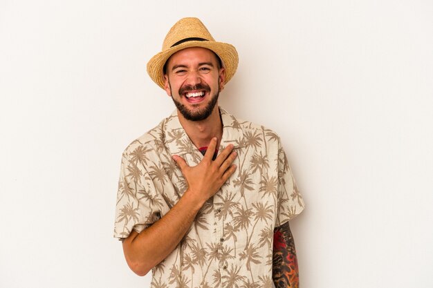 Young caucasian man with tattoos wearing summer clothes isolated on white background  laughs out loudly keeping hand on chest.