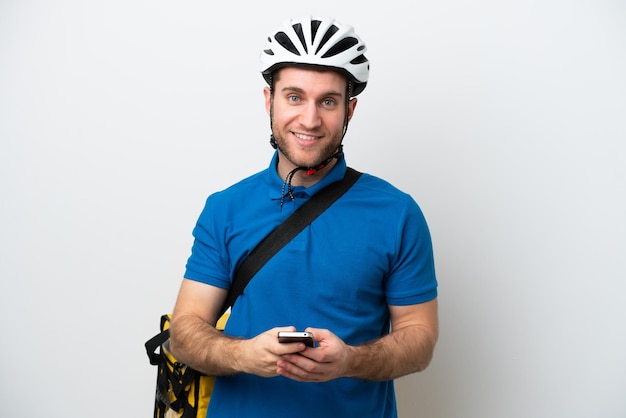 Young caucasian man with thermal backpack isolated on white background sending a message with the mobile