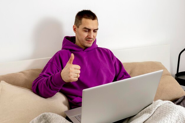 보라색 까마귀를 입은 백인 젊은이가 침대에 앉아 노트북 컴퓨터를 들고 있습니다. 노트북을 사용하여 온라인으로 공부하고 친구에게 전화하는 남자. 원격 교육, 화상 통화. 긍정적인 기호입니다.