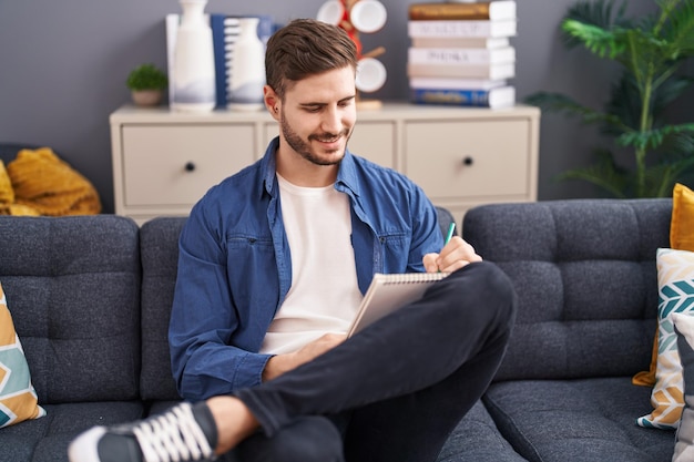 Young caucasian man writing on notebook sitting on sofa at home