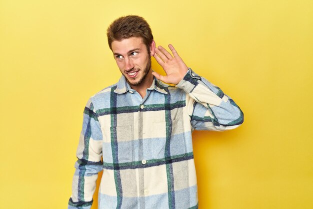 Young Caucasian man on a yellow studio background trying to listening a gossip