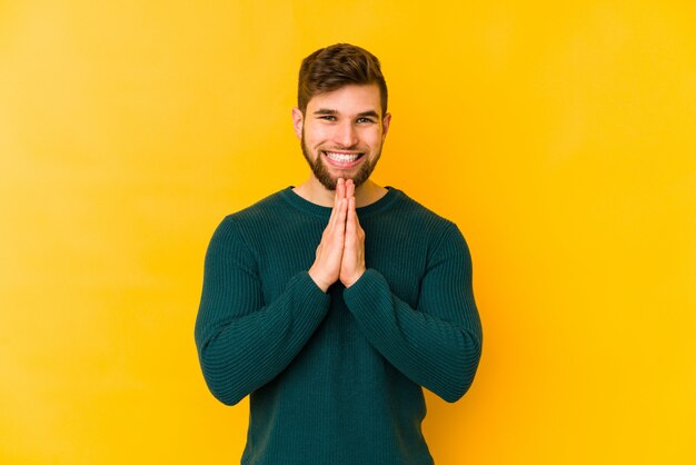 Young caucasian man on yellow wall holding hands in pray near mouth, feels confident.