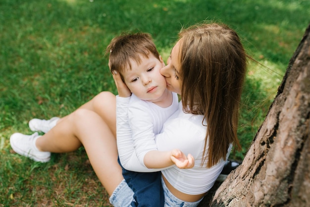 Young Caucasian mother kisses her threeyearold son on the cheek while sitting in the park near a tree on the grass in summer