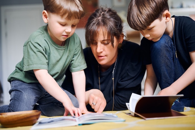 Young caucasian mother spending time at home with sons and reading books on the floor. Happy parent playing with preshool children. Home education concept