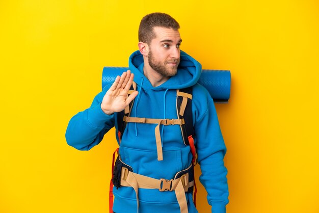 Foto giovane uomo caucasico dell'alpinista con un grande zaino isolato sulla parete gialla che fa gesto di arresto e deluso
