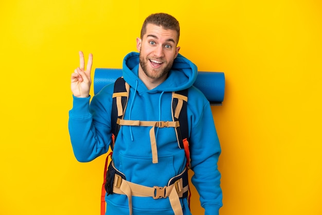 Young caucasian mountaineer man with a big backpack isolated on yellow wall smiling and showing victory sign