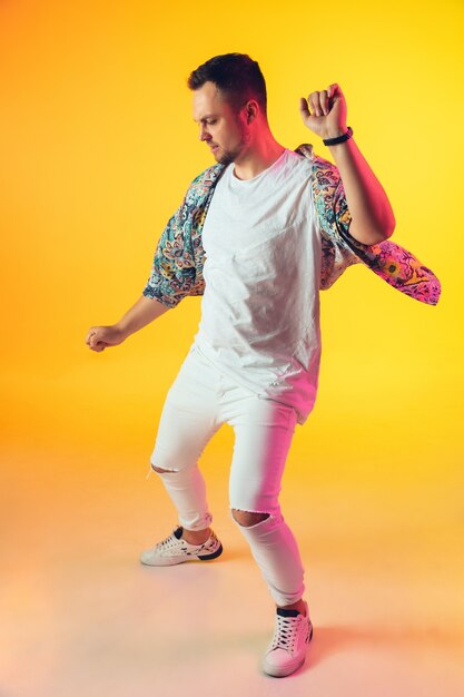 Photo young caucasian musician in casual dancing on gradient yellow background in neon light. concept of music, hobby, festival. joyful party host, dj, stand upper, dancer. colorful portrait of artist.