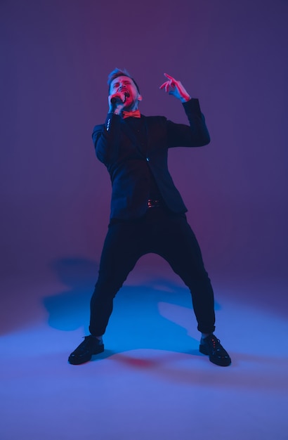 Young caucasian musician, performer singing, dancing in neon light on gradient background