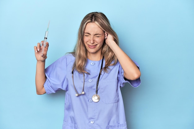Молодая белая медсестра в синей студии, прикрывающая уши руками.