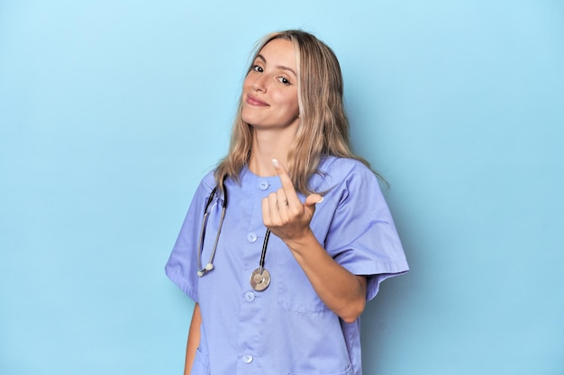 Young caucasian nurse in blue studio pointing with finger at you as if inviting come closer