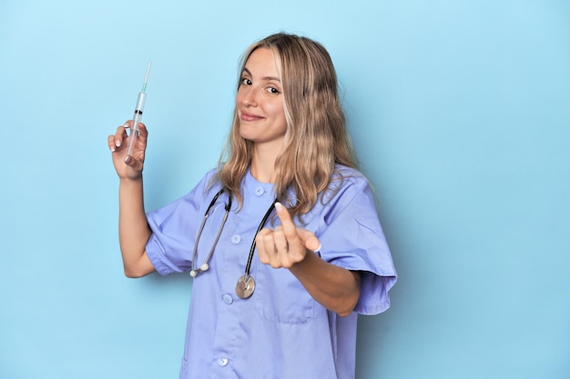 Foto giovane infermiera caucasica nello studio blu che ti indica con il dito come se ti invitasse ad avvicinarti