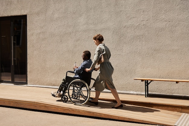 Young Caucasian nurse in dress pushing wheelchair with disabled man on ramp while he drinking refreshing cocktail