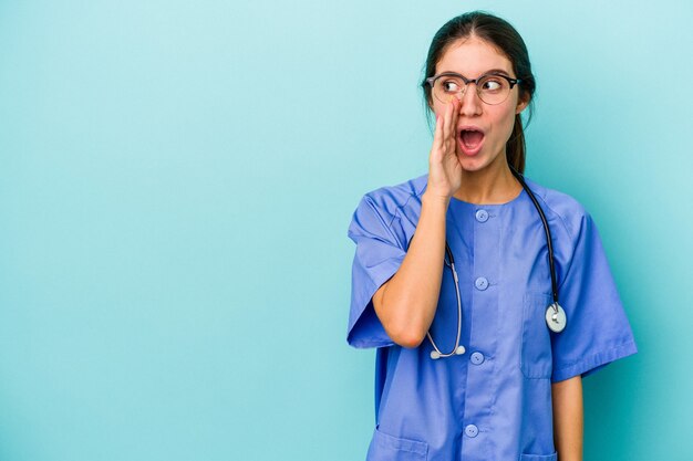 Young caucasian nurse isolated on blue background is saying a secret hot braking news and looking aside