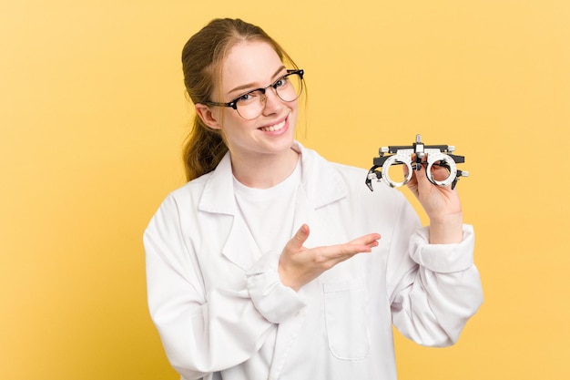 Giovane donna oculista caucasica che tiene un bicchiere di optometria isolato su sfondo giallo