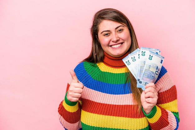 Young caucasian overweight woman holding banknotes isolated on pink background smiling and raising thumb up