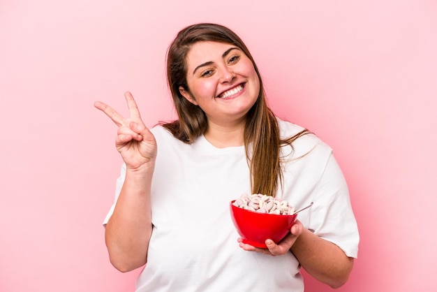 Young caucasian overweight woman holding a bowl of cereals isolated on pink background joyful and carefree showing a peace symbol with fingers