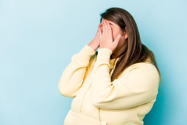 Young caucasian overweight woman isolated on blue background afraid covering eyes with hands.