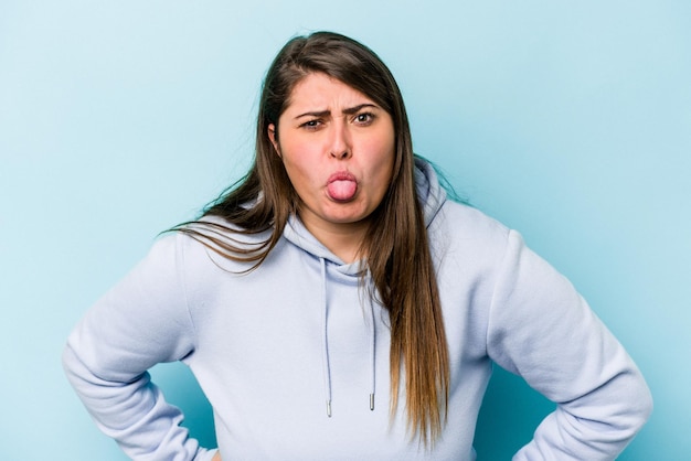 Young caucasian overweight woman isolated on blue background funny and friendly sticking out tongue.