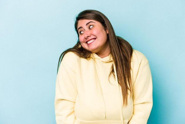 Young caucasian overweight woman isolated on blue background laughs and closes eyes, feels relaxed and happy.