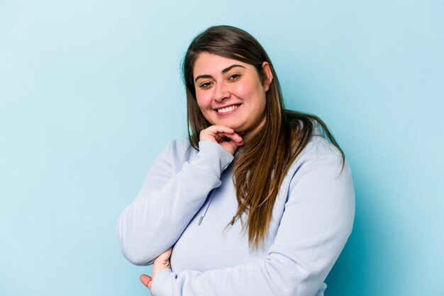 Young caucasian overweight woman isolated on blue background smiling happy and confident, touching chin with hand.